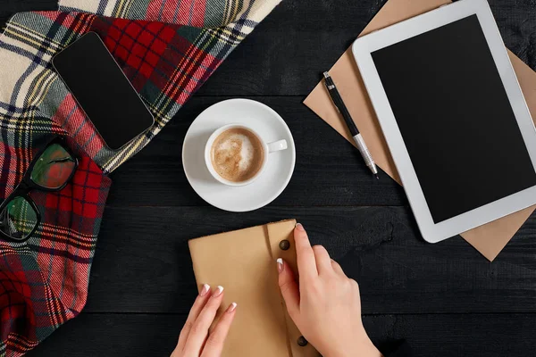 Молода жінка правою рукою пише на порожньому блокноті на дерев'яному столі з чашкою кави, смартфоном і окулярами поруч в ранковий час — стокове фото