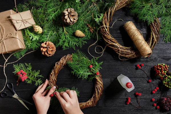 Visão superior de mãos femininas fazer uma grinalda de Natal. Presentes e pergaminhos embalados, ramos de abeto e ferramentas em mesa de madeira . — Fotografia de Stock