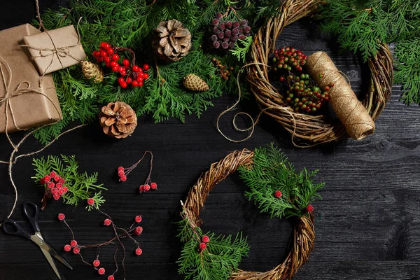 Faça uma coroa de Natal com suas próprias mãos. Ramo de abeto, grinalda de Natal e presentes em um fundo de madeira preta — Fotografia de Stock