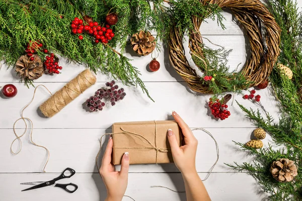 Вид сверху женских рук, завернутых в новогодний подарок. Упакованные подарки и свитки, еловые ветви и инструменты на деревянном столе . — стоковое фото