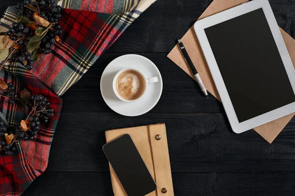 Вид зверху смартфон, планшет, шарф у клітці, окуляри з блокнотом та чашкою латте кави на чорному дерев'яному фоні . — стокове фото