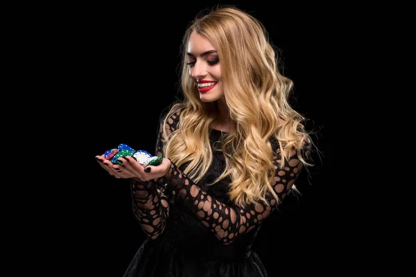 Elegant blonde in a black dress, casino player holding a handful of chips on black background