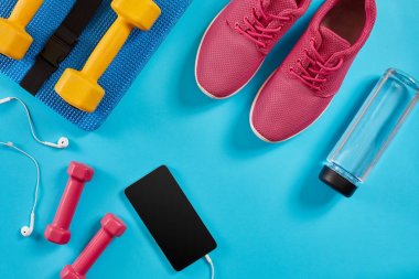 Spor ayakkabı, halter ve mavi arka plan üstünde hareket eden telefon. Üstten Görünüm. Fitness, spor ve sağlıklı yaşam konsepti.