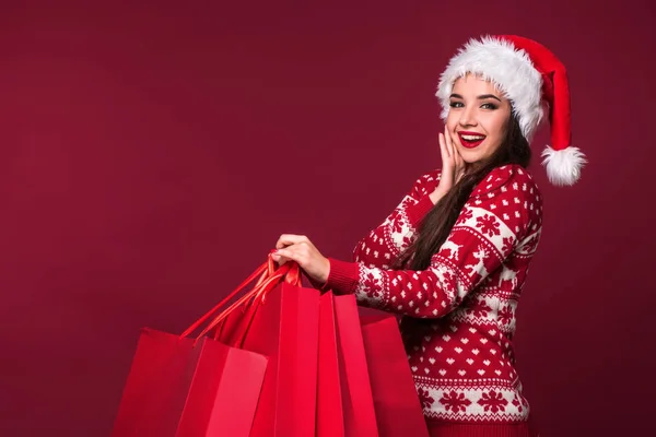 Young pretty woman or girl with long beautiful hair in Santa hat and New Years dress holding red package on red studio background — Stock Photo, Image