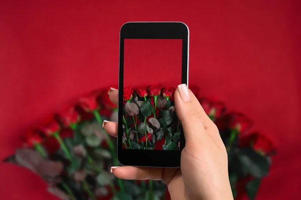 Closeup of woman is preparing to make a photo with a mobile phone. Bouquet of roses on a red background — Stock Photo, Image