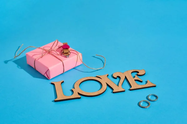 Puinen kirjoitus Rakkaus, lahjapakkaus ja sormus sinisellä taustalla. Avioehdotus. Käsite Ystävänpäivä — kuvapankkivalokuva