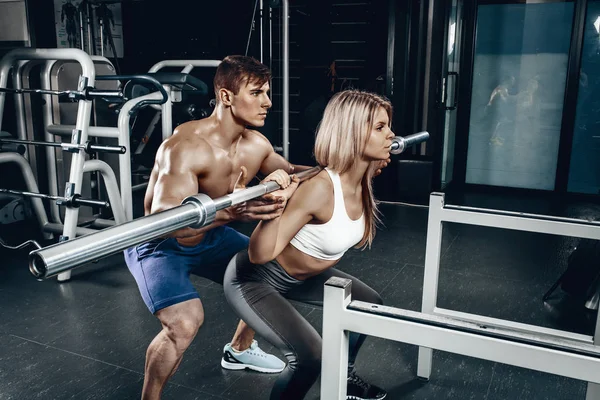 Personal trainer helping a young woman lift a barbell while working out in a gym