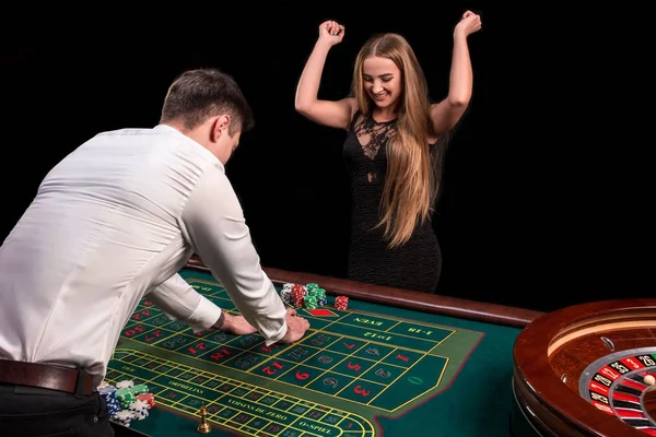 Un primer plano en la parte posterior del crupier en una camisa blanca, imagen de la mesa de casino verde con ruleta y fichas, una mujer rica apostando de juego en el fondo — Foto de Stock