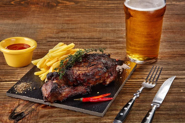 Bier wird in Glas mit Gourmet-Steak und Pommes frites auf Holzgrund gegossen. — Stockfoto