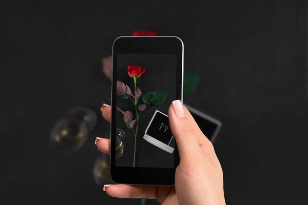 La mano femenina con un smartphone hace una foto una rosa roja oscura y una caja con pendientes sobre fondo negro . — Foto de Stock