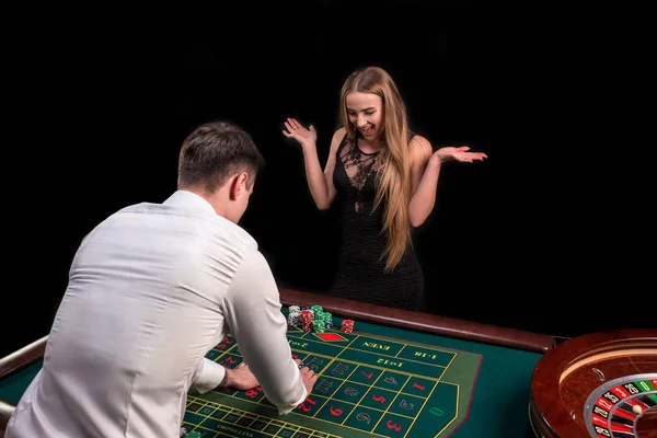 Un primer plano en la parte posterior del crupier en una camisa blanca, imagen de la mesa de casino verde con ruleta y fichas, una mujer rica apostando de juego en el fondo —  Fotos de Stock