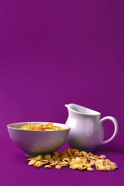 Cuenco con hojuelas de maíz, jarra de leche sobre fondo morado. oncept de un desayuno saludable —  Fotos de Stock