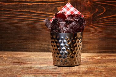 Dried peppered beef jerky cut in strips in a metal bucket on wooden background. Serving the dish in a restaurant clipart
