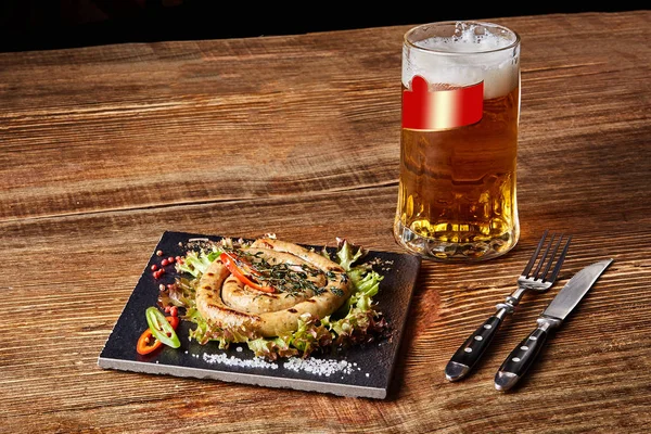 Hausgemachte Wurst mit Knoblauch und Gewürzen auf einem Schieferteller, ein Glas Bier hinter dem Teller auf einem Holztisch — Stockfoto