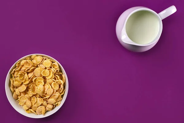 Cuenco con hojuelas de maíz, jarra de leche sobre fondo púrpura, vista superior —  Fotos de Stock