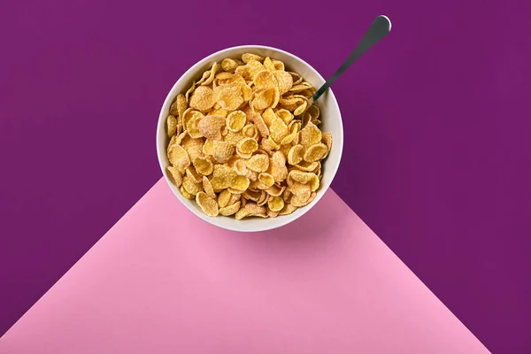 Cuenco con hojuelas de maíz y cuchara sobre fondo púrpura y rosa, vista superior —  Fotos de Stock