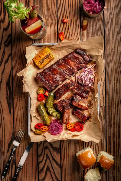 Grilled pork ribs served with grilled corn, salat, bbq sauce, salt pepper and cucumber on parchment paper on a wooden table. Top View — Stock Photo, Image