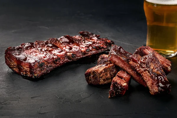 Pork ribs in barbecue sauce and a glass of beer on a black slate dish. A great snack to beer on a dark stone background. Top view with copy space — Stock Photo, Image