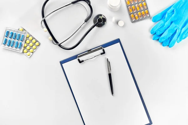 Equipamento médico: comprimidos, luvas azuis, termômetro e estetoscópio, branco em branco com uma caneta sobre fundo branco. Vista superior. Espaço de cópia — Fotografia de Stock