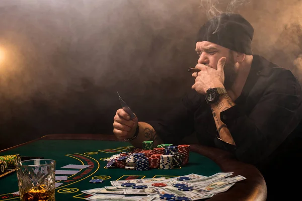 Skäggig man med cigarr och glas som sitter vid pokerbordet i ett kasino. Hasardspel, spelkort och roulette. — Stockfoto