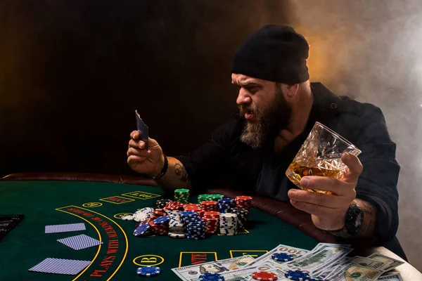 Homme barbu avec cigare et verre assis à la table de poker dans un casino. Jeux d'argent, cartes à jouer et roulette . — Photo