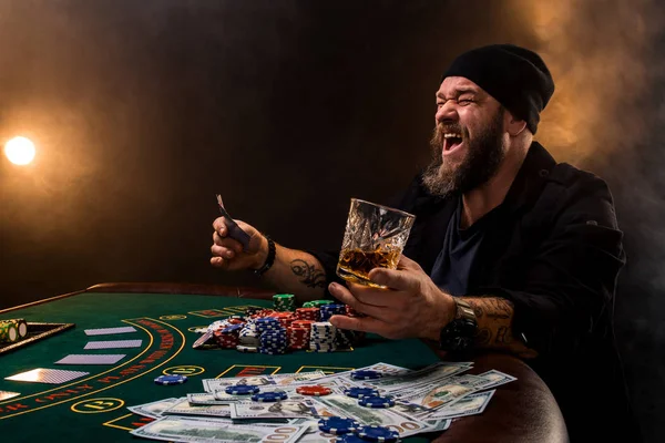 Skäggig man med cigarr och glas som sitter vid pokerbordet i ett kasino. Hasardspel, spelkort och roulette. — Stockfoto