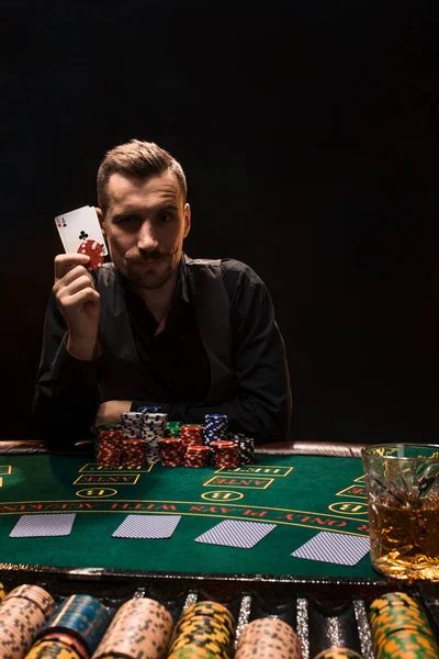 Guapo jugador de póquer con dos ases en sus manos y fichas sentado en la mesa de póquer sobre fondo negro —  Fotos de Stock