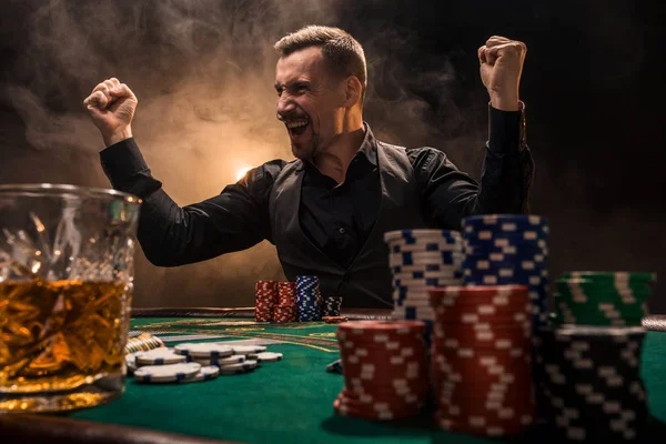 Joven hombre guapo sentado detrás de la mesa de póquer con cartas y fichas — Foto de Stock