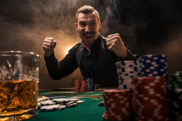 Joven hombre guapo sentado detrás de la mesa de póquer con cartas y fichas — Foto de Stock