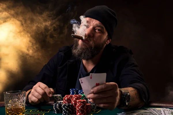 Skäggig man med cigarr och glas som sitter vid pokerbordet i ett kasino. Hasardspel, spelkort och roulette. — Stockfoto