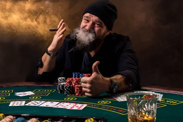 Vousatý muž s doutníkem a skla u stolu pokeru v kasinu. Hazardní hry, hrací karty a ruleta. — Stock fotografie