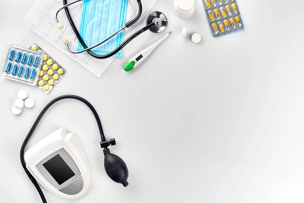Tonômetro elétrico branco com estetoscópio, pílulas multicoloridas, máscara e termômetro na mesa branca. Vista superior — Fotografia de Stock