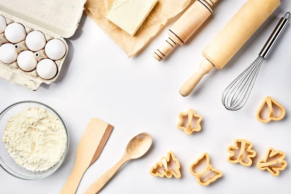 Bowl with wheat flour, rolling pin, whisk, eggs, butter, cookie cutters. Top view on a white table with a copy space