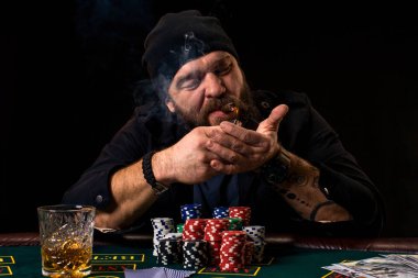 Bearded man with cigar and glass sitting at poker table in a casino. Gambling, playing cards and roulette. clipart