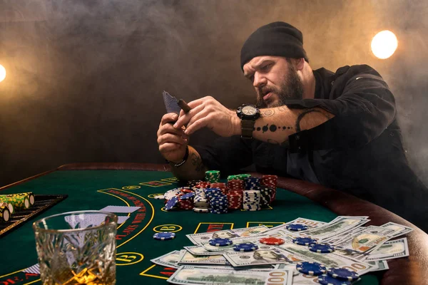 Hombre barbudo con cigarro y vidrio sentado en la mesa de póquer en un casino. Apuestas, cartas y ruleta . — Foto de Stock