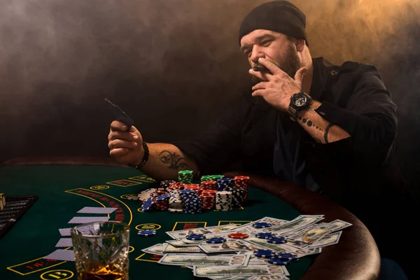 Bearded man with cigar and glass sitting at poker table in a casino. Gambling, playing cards and roulette.