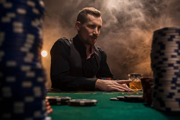 Joven hombre guapo sentado detrás de la mesa de póquer con cartas y fichas — Foto de Stock