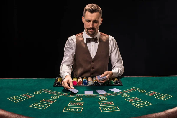 Portrét krupiér drží hrací karty, hracích známek na stole. Černé pozadí — Stock fotografie