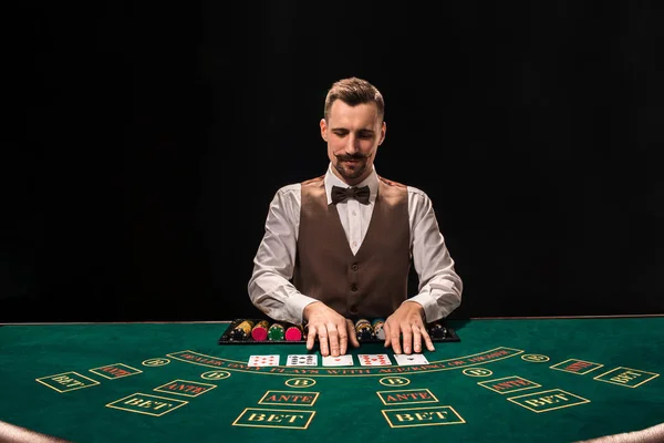 Portrait of a croupier is holding playing cards, gambling chips on table. Black background