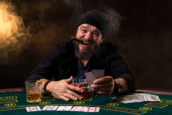 Skäggig man med cigarr och glas som sitter vid pokerbordet i ett kasino. Hasardspel, spelkort och roulette. — Stockfoto