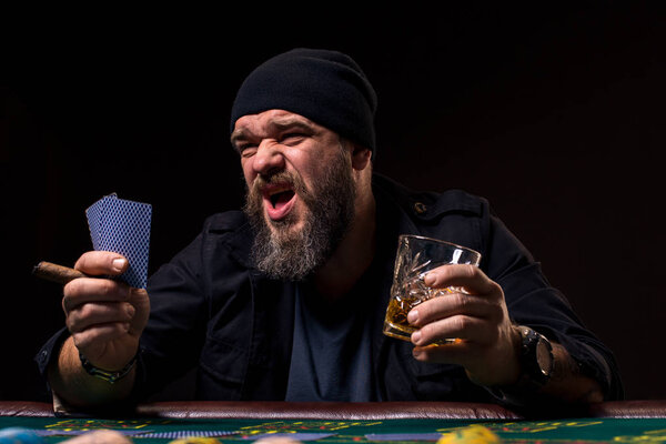 Bearded man with cigar and glass sitting at poker table and screaming isolated on black