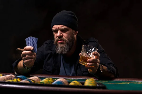 Allvarlig skäggig man med cigarr och glas som sitter vid pokerbordet och skrikande isolerade på svart — Stockfoto