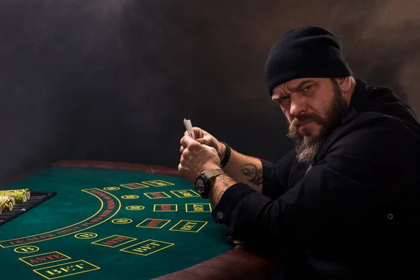 Σοβαρή γενειοφόρος άνδρας κάθεται στο τραπέζι του πόκερ και κρατώντας κάρτες που απομονώνονται σε μαύρο. Χώρο αντίγραφο — Φωτογραφία Αρχείου