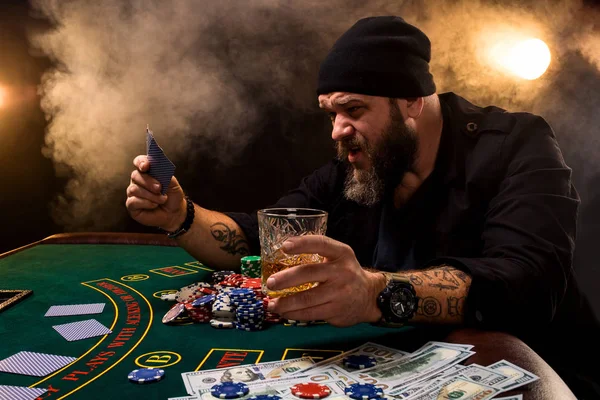 Vousatý muž s doutníkem a skla u stolu pokeru v kasinu. Hazardní hry, hrací karty a ruleta. — Stock fotografie