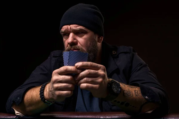 Serious bearded man sitting at poker table and holding cards isolated on black Stock Picture