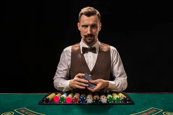 Portret krupierka trzyma karty do gry, hazard żetonów na stole. Czarne tło — Zdjęcie stockowe