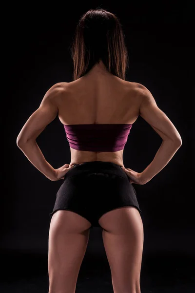 Jovem bela mulher de cabelos escuros em sutiã esportivo roxo escuro e shorts curtos pretos sedutoras posando com as costas contra um fundo preto — Fotografia de Stock
