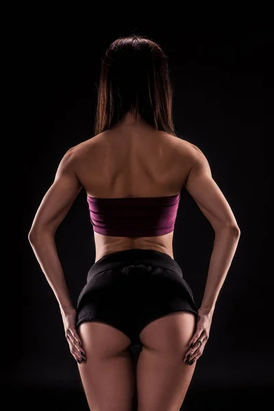 Jeune belle femme aux cheveux bruns en soutien-gorge de sport violet foncé et short noir posant séduisante avec le dos sur un fond noir — Photo