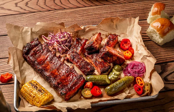 Grilled pork ribs served with grilled corn, salat, bbq sauce, salt pepper and cucumber on parchment paper on a wooden table. Top View — Stock Photo, Image