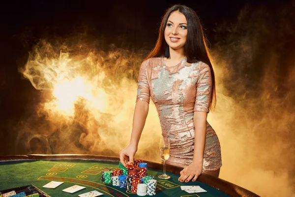 Wanita muda cantik berpakaian malam berdiri di dekat meja poker dengan segelas sampanye — Stok Foto